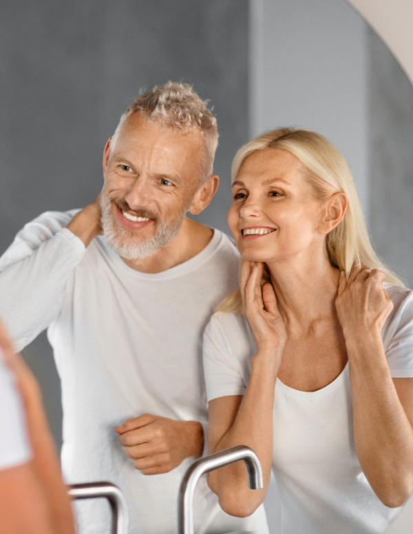 Older couple smiling and looking into a mirror, both dressed in white shirts, touching their necks.