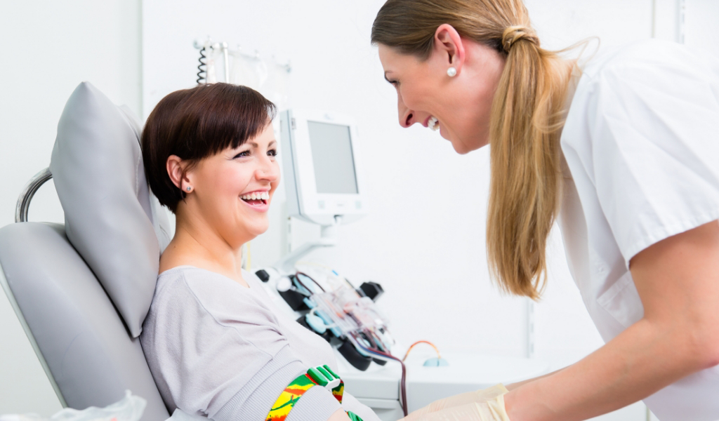 A patient sits in a medical chair, smiling, while a healthcare professional assists her. Medical equipment is visible in the background.