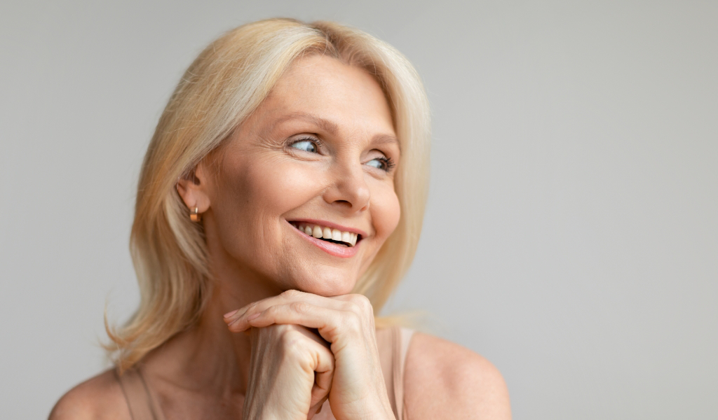 A smiling woman with blonde hair, in a light-colored top, rests her chin on her hands and looks to the side against a plain background.