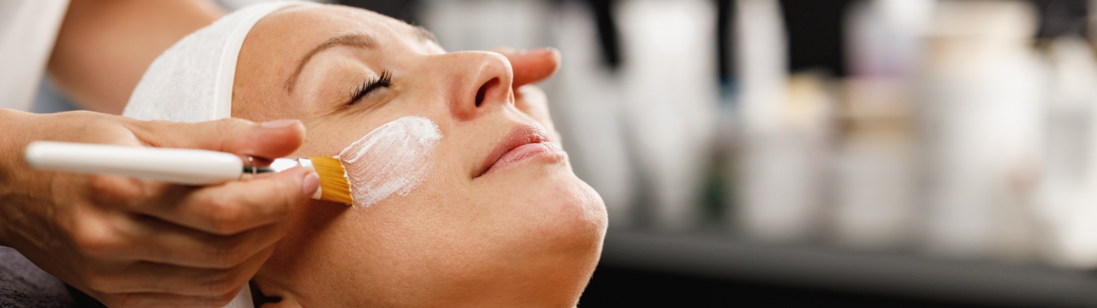 Person receiving a facial treatment with a brush applying a white mask while lying down with eyes closed.