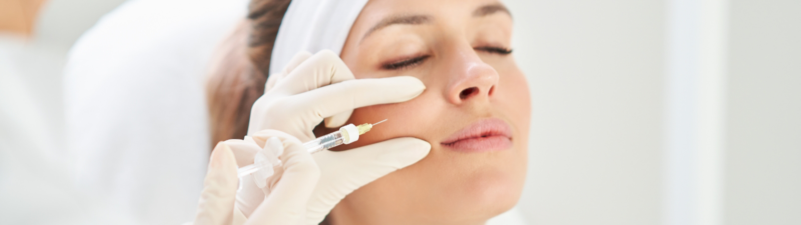 A person receiving a cosmetic injection on the side of their face while lying down, administered by another individual wearing white gloves.