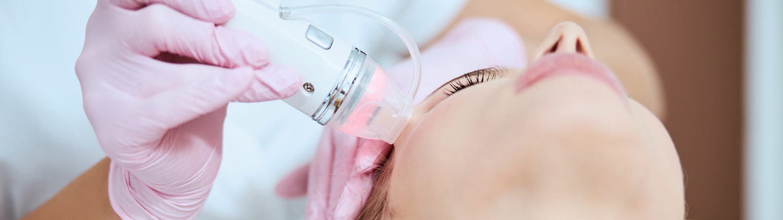 A person lying down receives a facial treatment with a handheld device operated by a professional wearing pink gloves.