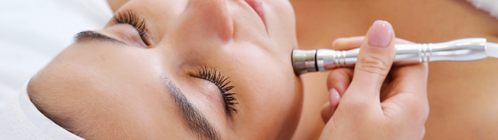 A person lying down with closed eyes receives a facial treatment using a metal device applied to their chin.