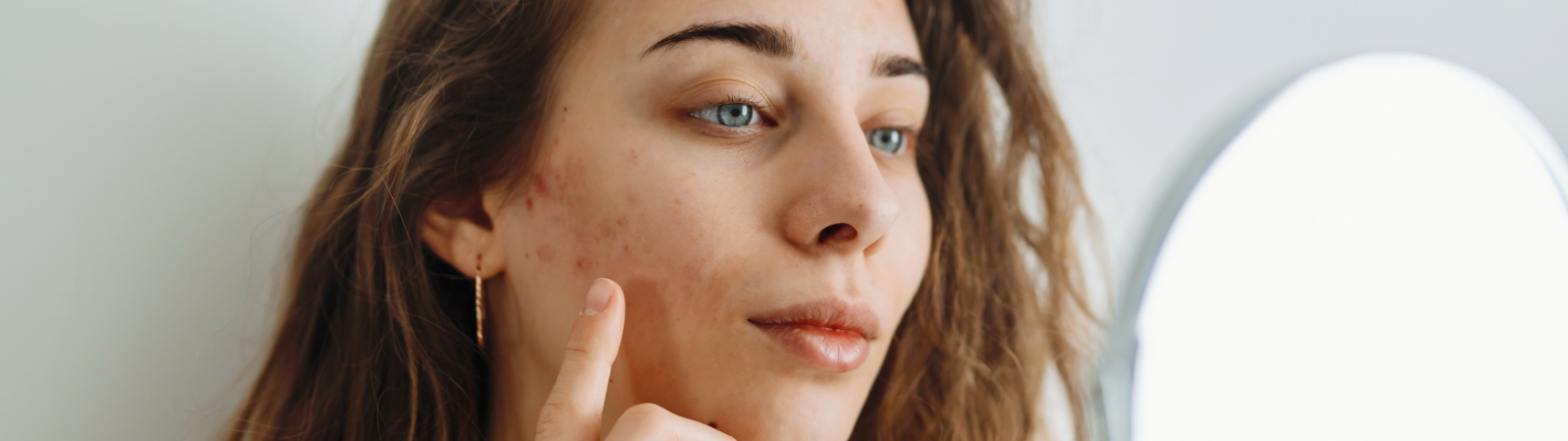 A woman with long hair examines her face in a mirror, pointing at a small blemish on her cheek.