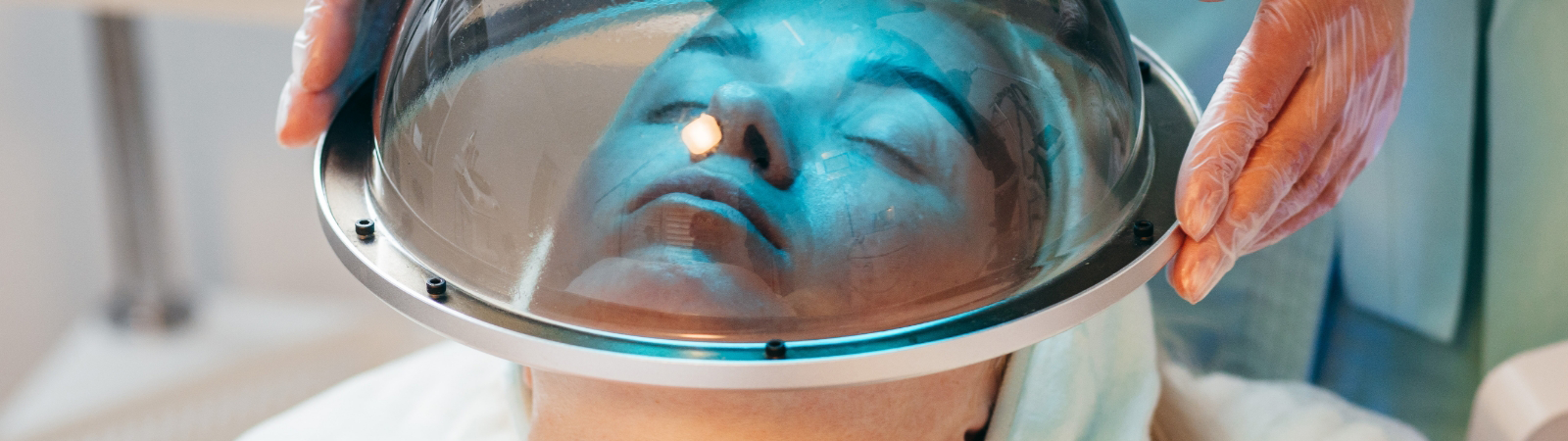 A person lies with their face under a circular glass dome, while gloved hands hold the dome in place, suggesting a skincare or facial treatment procedure.