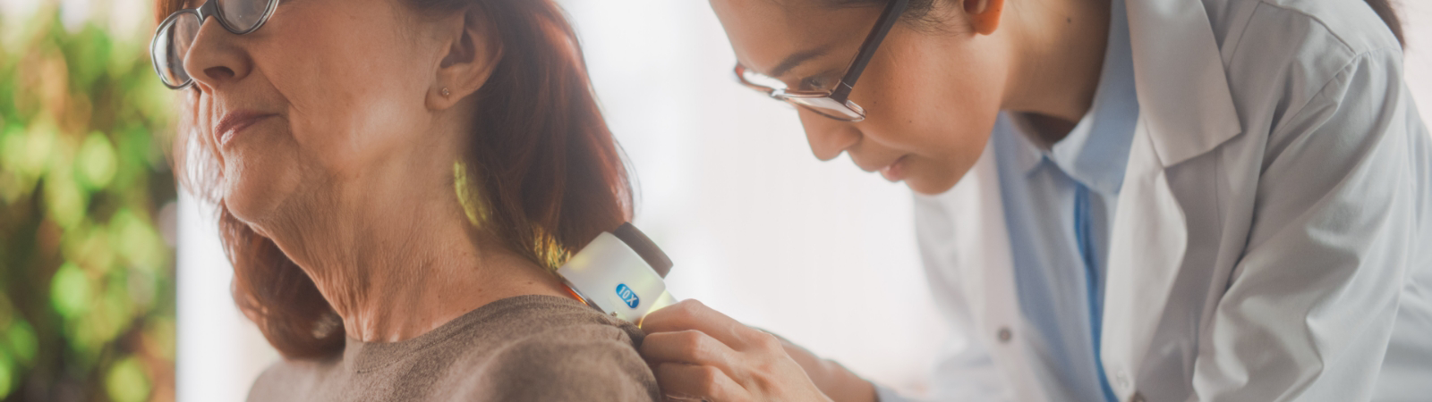 A healthcare professional examines the skin on an older woman's upper back using a medical instrument. Both are wearing glasses.