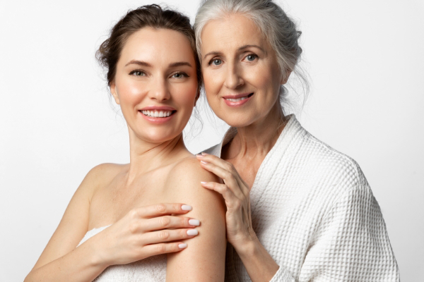 A younger woman and an older woman stand closely together, smiling at the camera. The younger woman is wrapped in a towel, and the older woman is wearing a white robe. They both have light skin and brown hair.